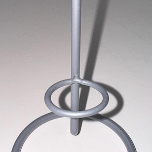 Puede incluir: Un soporte decorativo de metal plateado con una base circular, un anillo central y un poste vertical que termina en dos brazos curvos. El soporte está diseñado para sostener algo.