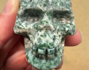 May include: A carved skull figurine made of green and white speckled stone. The skull has detailed features, including eye sockets, a nose cavity, and a full set of teeth. The stone has a natural, textured appearance.