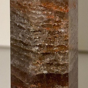 May include: A tall, rectangular crystal point with a faceted top. The crystal features bands of brown, white, and reddish-brown hues, creating a layered appearance. The crystal is a natural stone.