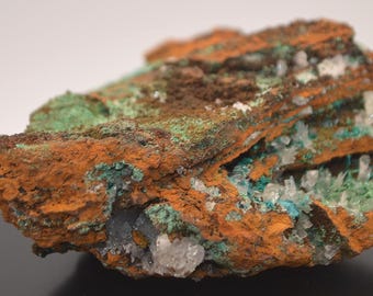 May include: A close-up of a mineral specimen with a rough, layered texture. The rock features vibrant orange, green, and white crystalline formations. The colors and textures create a striking visual contrast, highlighting the natural beauty of the mineral.
