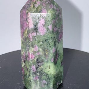 Könnte beinhalten: Ein facettierter, polierter Edelstein mit grüner Grundfarbe und rosa Einschlüssen. Der Kristall ist rechteckig mit einer spitzen Spitze. Der Stein wird auf einer schwarzen Oberfläche präsentiert.