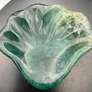 Op de afbeelding: Een doorschijnende groene glazen schaal met een handvormig ontwerp. De schaal heeft een geschulpte rand en zichtbare scheuren, met een lichter groen en geel getint gebied aan één kant. De schaal staat op een donkere ondergrond.