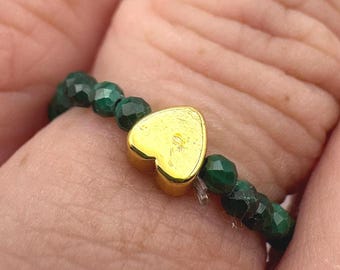Peut inclure: Une bague de perles avec des perles facettées vert foncé et un charm en forme de cœur doré. La bague est portée au doigt, mettant en valeur le contraste entre les perles vertes et le cœur doré.