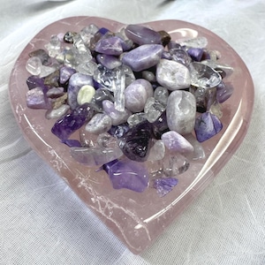 May include: A heart-shaped, light pink dish filled with an assortment of polished gemstones. The gemstones are primarily shades of purple, lavender, and clear, with some having a translucent quality. The dish is sitting on a white surface.