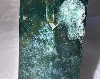May include: A polished, rectangular moss agate crystal. The stone is primarily green with white and translucent inclusions. The crystal is cut into a tall, obelisk shape, with a flat base and a pointed top.