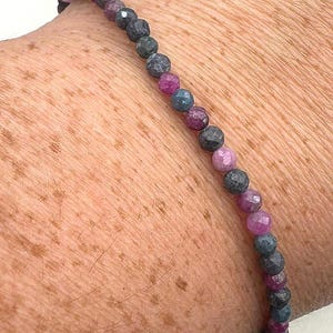 Op de afbeelding: Een delicate kralenarmband met een reeks kleine, gefacetteerde edelstenen in tinten roze, blauw en grijs. De armband is geregen op een dun, flexibel koord en is ontworpen om om de pols te worden gedragen.
