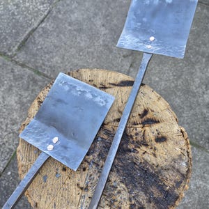 May include: Two metal pizza peels with square blades and long handles rest on a wooden tree stump. The blades have a hammered texture and are attached to the handles with rivets. The metal has a dark, slightly bluish hue.