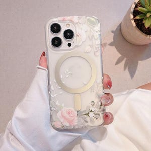 Op de afbeelding: Transparante telefoonhoes met een bloemenpatroon van roze rozen en witte accenten. De hoes heeft een goudkleurige magnetische ring en een camera-uitsparing. Het ontwerp is delicaat en vrouwelijk.