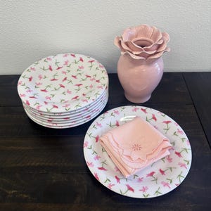 May include: A stack of white ceramic plates with a pink floral pattern, a pink vase with a flower-shaped top, and a plate with a folded pink napkin. The plates and napkin have a matching floral design.