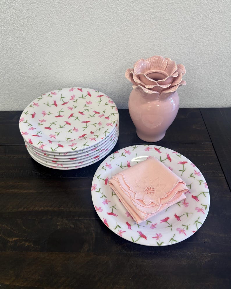 May include: A stack of white ceramic plates with a pink floral pattern, a pink vase with a flower-shaped top, and a plate with a folded pink napkin. The plates and napkin have a matching floral design.
