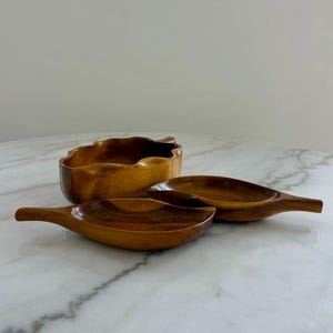 May include: A set of three wooden serving bowls. The set includes two leaf-shaped bowls with handles and a larger, scalloped-edge bowl. The bowls are made of a warm-toned wood and are arranged on a marble surface.