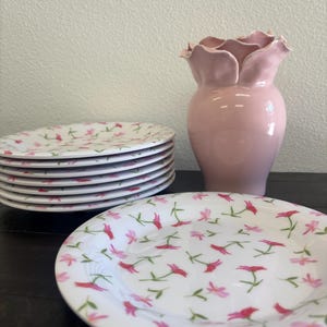 May include: A stack of white ceramic plates with a pink floral pattern, next to a pink vase with a ruffled top. The plates are stacked on a dark surface, and the vase is a solid, soft pink color.