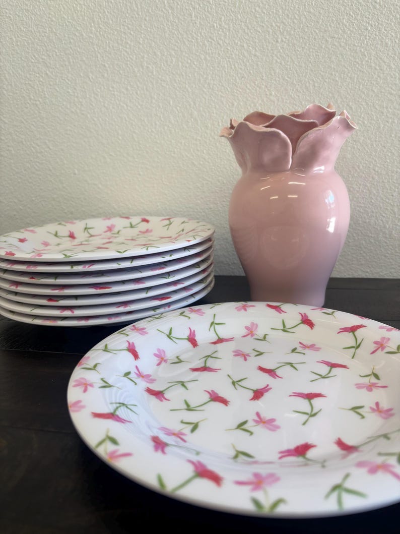 May include: A stack of white ceramic plates with a pink floral pattern, next to a pink vase with a ruffled top. The plates are stacked on a dark surface, and the vase is a solid, soft pink color.