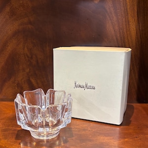 May include: A clear, faceted glass bowl with a unique, scalloped rim. The bowl sits on a wooden surface next to a box with the text "Neiman Marcus" printed on it. The bowl's design is reminiscent of a flower.