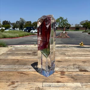 May include: A clear, rectangular resin sculpture with a large, faceted, dark red and green crystal embedded inside. The sculpture is displayed on a wooden surface, with a background of a parking lot and trees.