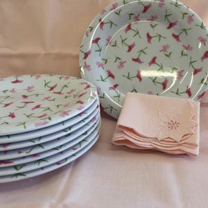 May include: A stack of white plates with a floral pattern of pink and red flowers and green stems. A matching bowl and a stack of pink napkins with a floral embroidered design are also present. The items are arranged on a pink surface.