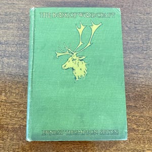 May include: A vintage book titled "The Book of Woodcraft" with a green cover. The cover features a yellow illustration of a deer with large antlers. The author's name, Ernest Thompson Seton, is printed at the bottom of the cover.