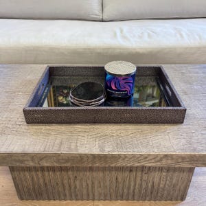 May include: A decorative tray with a mirrored base, holding a candle and coasters. The tray has a brown, textured exterior and handles. The candle has a silver lid and a blue, purple, and pink design. The coasters are dark gray.