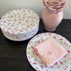May include: A stack of white ceramic plates with a floral pattern of pink and red flowers. A pink napkin with a floral design sits on a matching plate. A pink flower-shaped vase is also present.