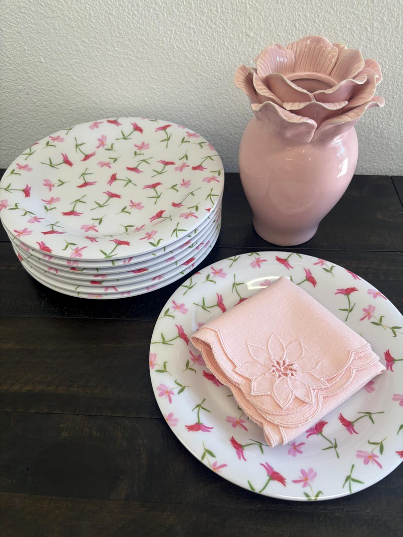 May include: A stack of white ceramic plates with a floral pattern of pink and red flowers. A pink napkin with a floral design sits on a matching plate. A pink flower-shaped vase is also present.