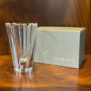 May include: A clear, fluted glass vase with a red and white label, next to a gray box with the text "Orrefors". The vase has a unique, textured design and is made of crystal. The box is rectangular and has the Orrefors logo.