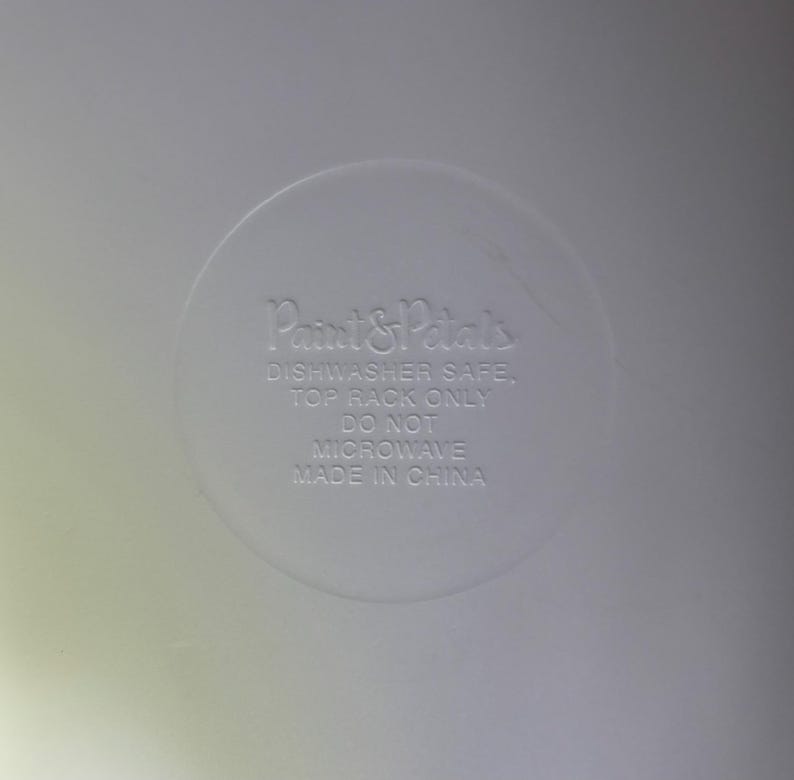 May include: A close-up of a white, circular design with the text "Paint & Petals" and care instructions: "Dishwasher Safe, Top Rack Only, Do Not Microwave, Made in China."