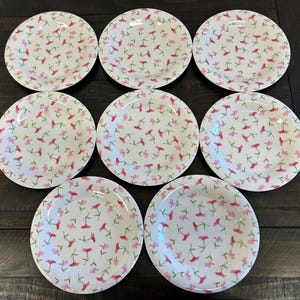 May include: A set of eight white ceramic plates with a floral pattern. The plates feature a repeating design of pink and red flowers with green stems. The plates are arranged on a dark wooden surface.