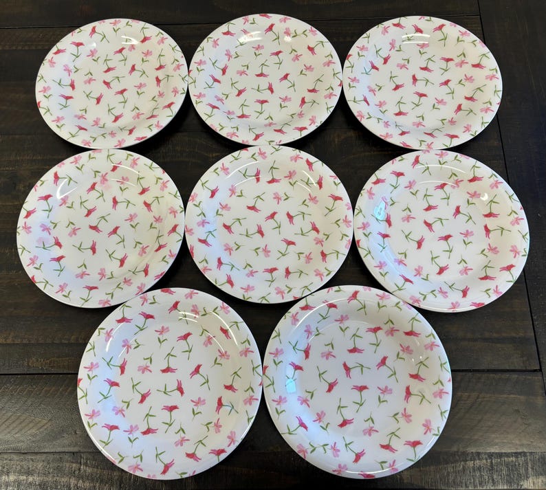 May include: A set of eight white ceramic plates with a floral pattern. The plates feature a repeating design of pink and red flowers with green stems. The plates are arranged on a dark wooden surface.