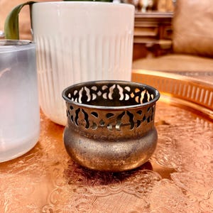 May include: A small, antique-style metal candle holder with a decorative cut-out design around the top. The candle holder has a tarnished, bronze-like finish. It sits on a copper-colored tray with an ornate pattern, alongside a white ceramic pot and a glass candle holder.