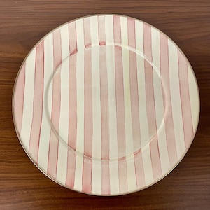 May include: A round, clear glass plate with a gold rim. The plate features a vertical striped pattern in alternating shades of pink and white. The plate is suitable for serving food.