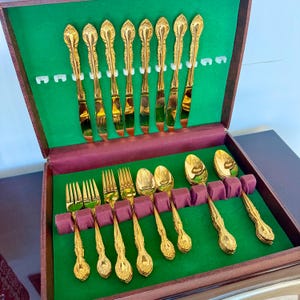 May include: A gold-toned flatware set housed in a dark wooden storage case with a green velvet interior. The set includes eight knives with ornate handles, four forks, two spoons, and two larger spoons, all featuring a matching decorative pattern.