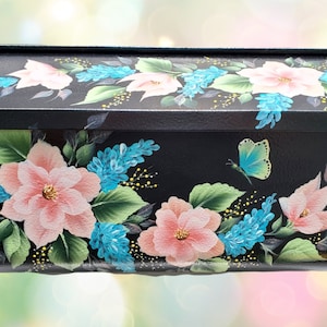 May include: Black mailbox with hand-painted floral design. The flowers are pink and blue with green leaves, and a light blue butterfly. The mailbox has a black roof and is rectangular in shape.