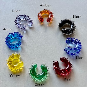 May include: A collection of colorful ear cuffs in a variety of colors, including aqua, blue, amber, black, green, lilac, red, and yellow. Each cuff is a unique, C-shaped design with a textured, beaded edge. The names of the colors are labeled on the image.