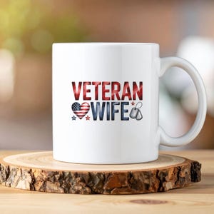 May include: White ceramic mug with the words "VETERAN WIFE" in red, white, and blue. The design includes a heart with an American flag pattern, stars, and dog tags. The mug sits on a wooden slice.