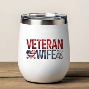 May include: White stemless wine tumbler with the words "VETERAN WIFE" in red and blue. The design includes a heart with an American flag pattern, stars, and dog tags. The tumbler has a silver rim and lid.
