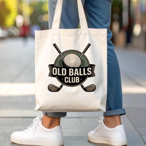 Puede incluir: Bolso tote blanco con un dise&ntilde;o tem&aacute;tico de golf. El dise&ntilde;o incluye una pelota de golf, palos de golf cruzados y el texto "OLD BALLS CLUB" en una pancarta. El bolso lo lleva una persona con vaqueros azules y zapatillas blancas.
