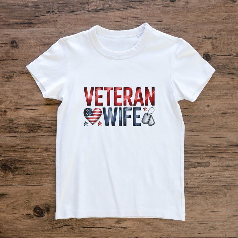 May include: White t-shirt with the words "VETERAN WIFE" in red, white, and blue. The design includes a heart with an American flag pattern, stars, and dog tags. The shirt is made of a soft material.
