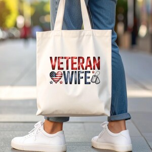 May include: A white tote bag with the words "VETERAN WIFE" in red, white, and blue. The design includes a heart with the American flag, stars, and dog tags. The bag has two white handles.