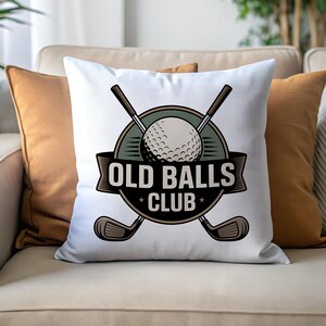 Puede incluir: Coj&iacute;n cuadrado blanco con un dise&ntilde;o tem&aacute;tico de golf. El dise&ntilde;o presenta una pelota de golf, palos de golf cruzados y el texto "OLD BALLS CLUB" en un emblema circular. El coj&iacute;n est&aacute; sobre un sof&aacute; beige con cojines naranjas.