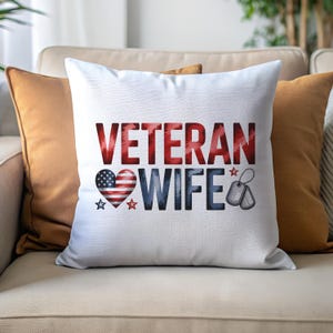 May include: White decorative pillow with the words "VETERAN WIFE" in red and blue. A heart-shaped American flag graphic is on the left, and dog tags are on the right. The pillow is on a beige couch with orange pillows.