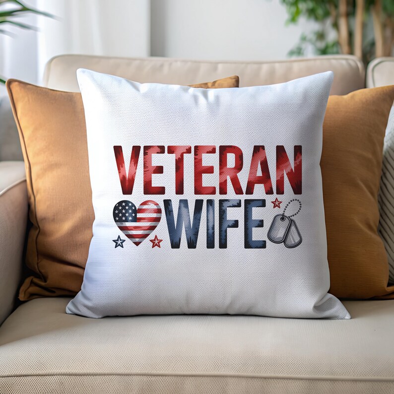 May include: White decorative pillow with the words "VETERAN WIFE" in red and blue. A heart-shaped American flag graphic is on the left, and dog tags are on the right. The pillow is on a beige couch with orange pillows.