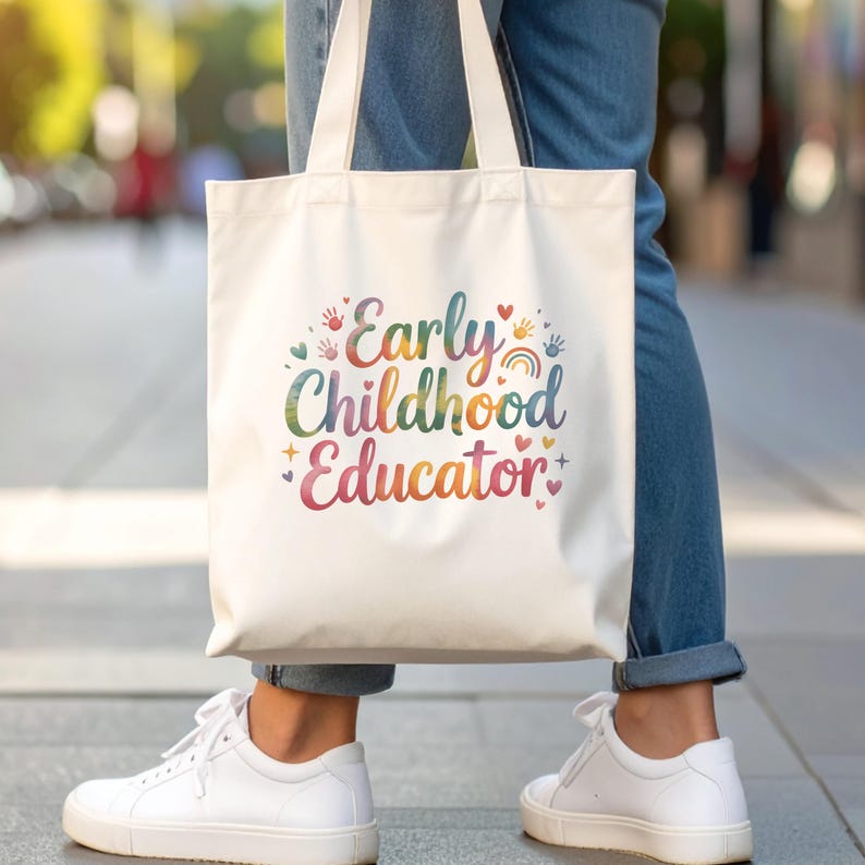 May include: A white tote bag featuring the words "Early Childhood Educator" in a rainbow-coloured design. The bag has white handles and is held by someone wearing blue jeans and white trainers.