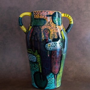 May include: A ceramic vase with two yellow handles featuring a glossy finish and a multi-colored abstract pattern. The surface displays hand-painted shapes in shades of green, orange, black, and blue, including squiggles, dots, and solid forms.