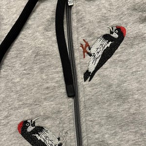 May include: Gray zip-up hoodie featuring embroidered woodpecker designs. The woodpeckers are black, white, and red, with detailed stitching. The hoodie has a black drawstring and a black zipper. A casual, comfortable garment.