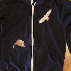 Könnte beinhalten: Ein marineblauer Kapuzenpullover mit Reißverschluss, weißem Reißverschluss und zwei Vordertaschen. Der Kapuzenpullover hat ein gesticktes Falken- und Maus-Design. Der Falke ist mit braunen und weißen Federn dargestellt, und die Maus ist braun.