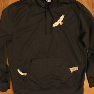 Puede incluir: Sudadera con capucha negra con bolsillo delantero y capucha con cordón. La sudadera presenta un diseño bordado de un halcón en vuelo en la parte superior izquierda del pecho. Detalles bordados adicionales están en el bolsillo.