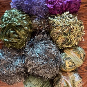 May include: A collection of yarn balls in various colors and textures. The yarns include shades of purple, green, and gray, with some having a fuzzy or ribbon-like appearance. The image showcases a variety of textures, from smooth to textured, suitable for crafting projects.