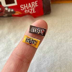 May include: Two miniature M&M's candy wrappers, one black and one yellow, rest on a fingertip. The black wrapper has a brown and white logo, while the yellow wrapper has a yellow and red logo. A red and brown M&M's bag is in the background.