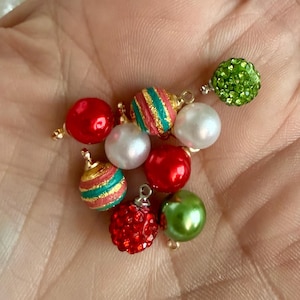 May include: A collection of small spherical charms shaped like holiday ornaments. The set includes red, white, and green glossy spheres, two spheres with red, green, and gold striped patterns, and two spheres covered in small red and green crystals.