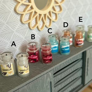 May include: A collection of small, glass jar candles with various scents. The candles are labeled with text such as "Vanilla", "Apple Cider", "Sea Breeze", and "Pumpkin". The jars are arranged on a gray cabinet, with a decorative mirror in the background.
